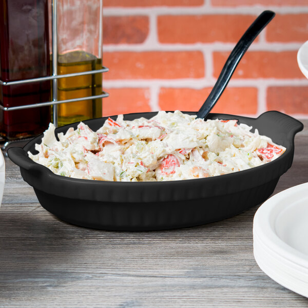A black Tablecraft shallow oval casserole dish filled with food on a table.
