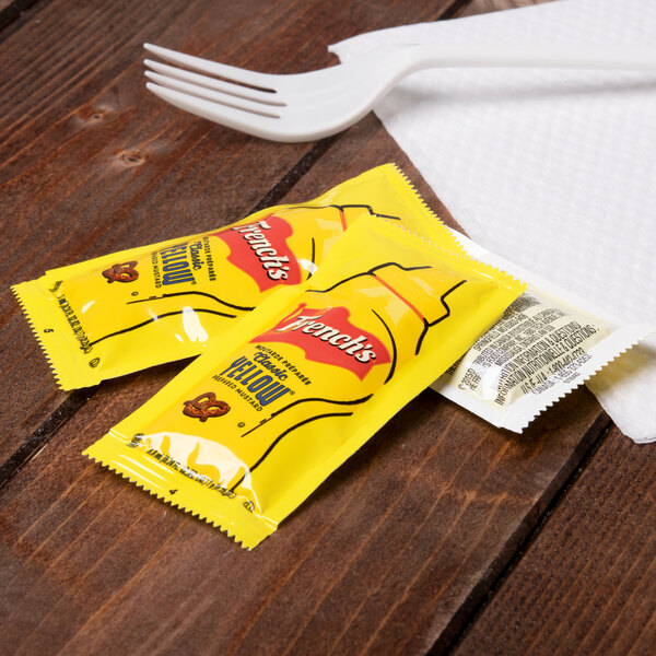 French's Classic Yellow Mustard packets next to a fork on a table with a spoon and napkin.