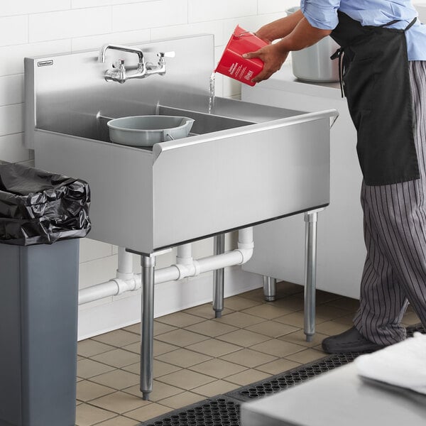 A woman in an apron standing in a school kitchen washing dishes in a Steelton stainless steel utility sink.