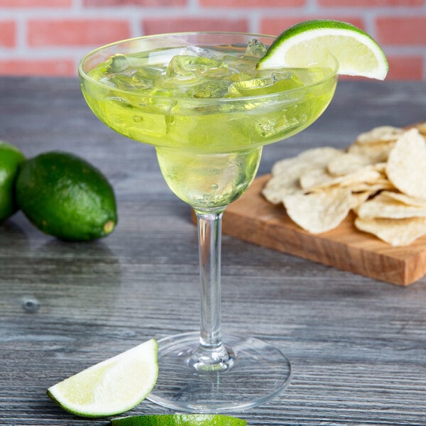 A Thunder Group plastic margarita glass filled with a drink and garnished with a lime wedge on a wooden table.