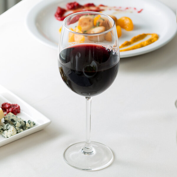 An Arcoroc Rutherford tall wine glass filled with red wine on a table with food.