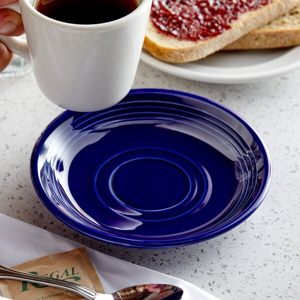 A hand holding a white cup of coffee over a Tuxton cobalt blue saucer.