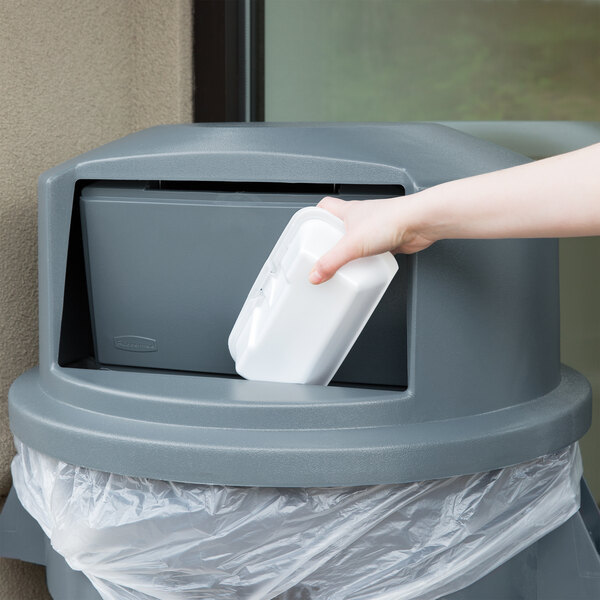 A hand throwing a white container into a grey trash can.