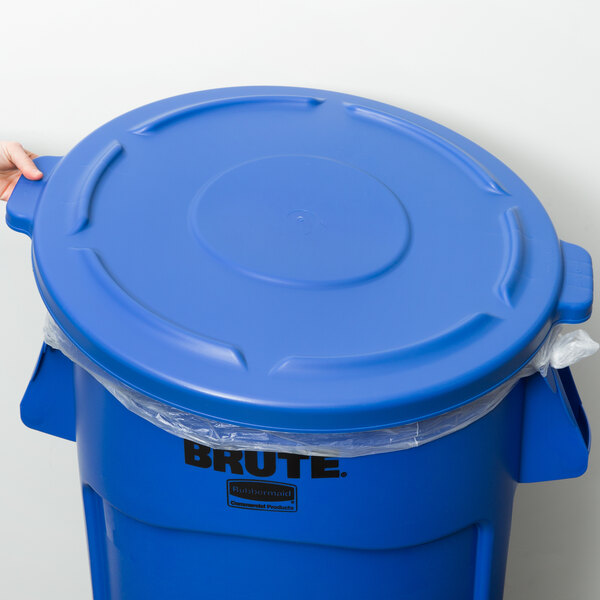 A hand placing a Rubbermaid BRUTE blue trash can lid on a blue Rubbermaid trash can.