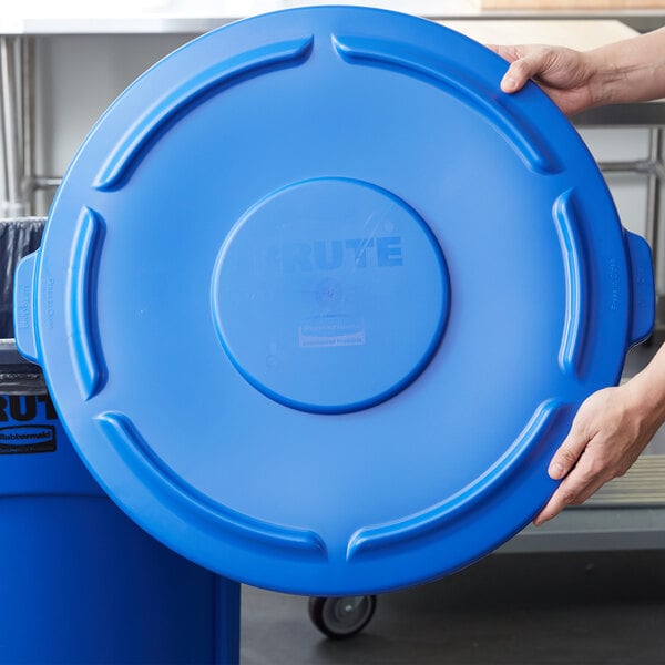 A person holding a blue plastic lid over a blue Rubbermaid BRUTE trash can.