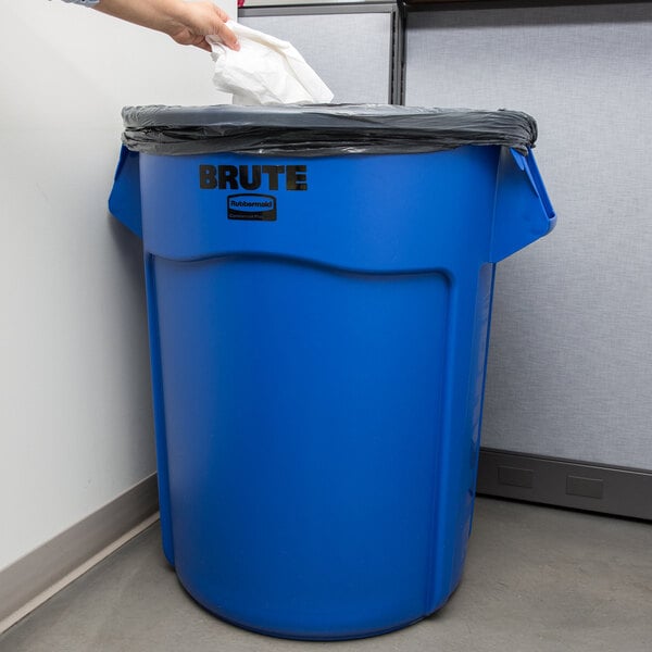 A hand putting a black plastic liner in a blue Rubbermaid BRUTE trash can.