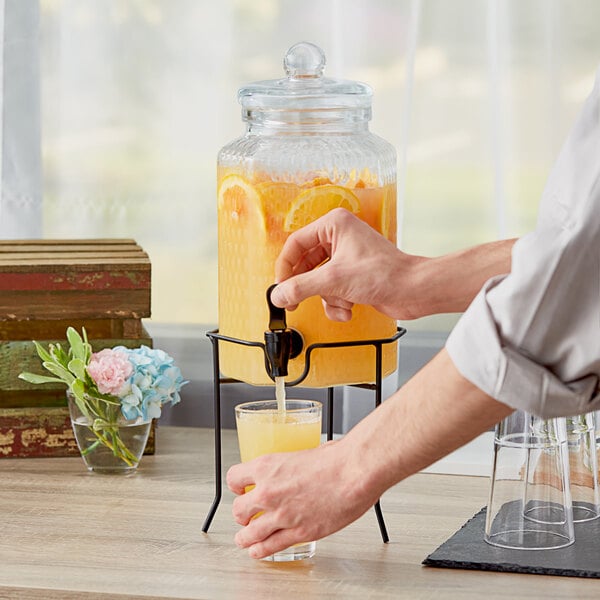 A hand pressing the Acopa glass beverage dispenser to pour orange juice into a glass.