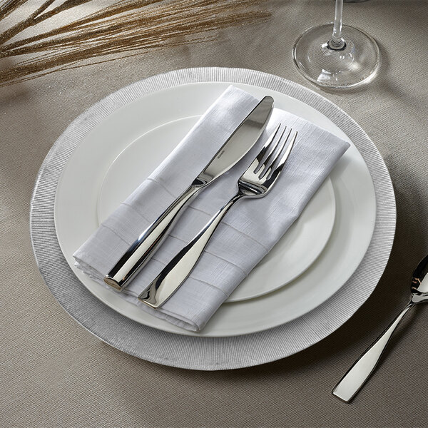 A white plate with a silver glass charger on it and silverware and a napkin.