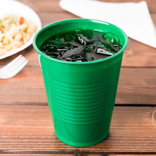 A Creative Converting emerald green plastic cup filled with ice on a wooden table with a plate of food.