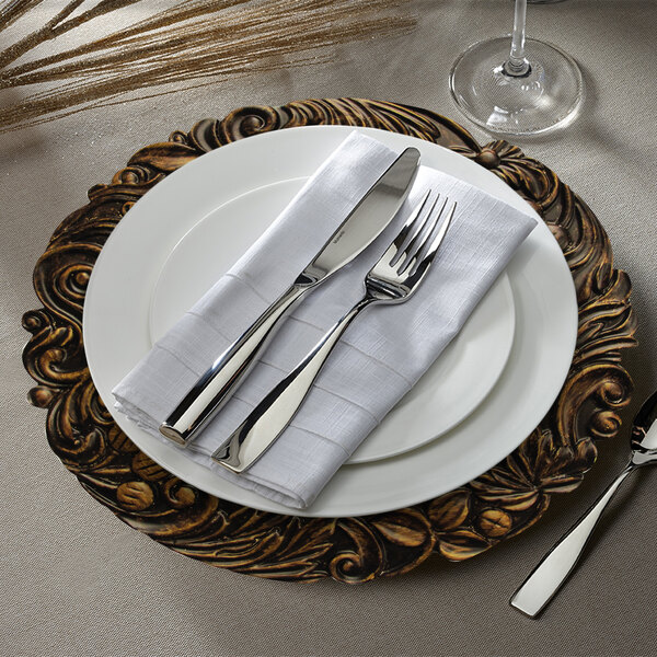 A dark oak Charge It by Jay plastic charger plate with silverware on a white napkin.