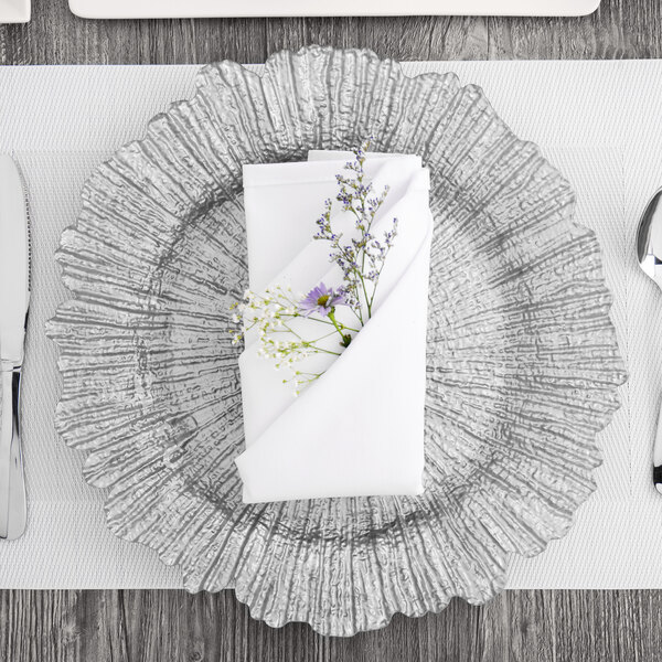 A Charge It by Jay silver glass charger plate with a white napkin and purple and white flowers on it.
