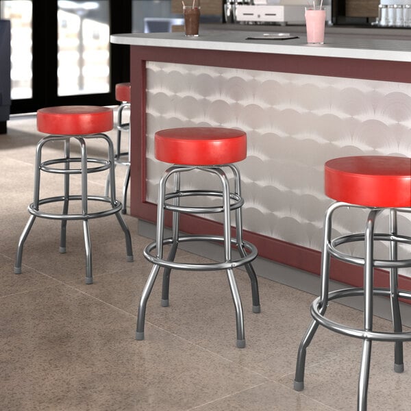 Three Lancaster Table & Seating red vinyl double ring swivel bar stools with metal legs at a bar counter.