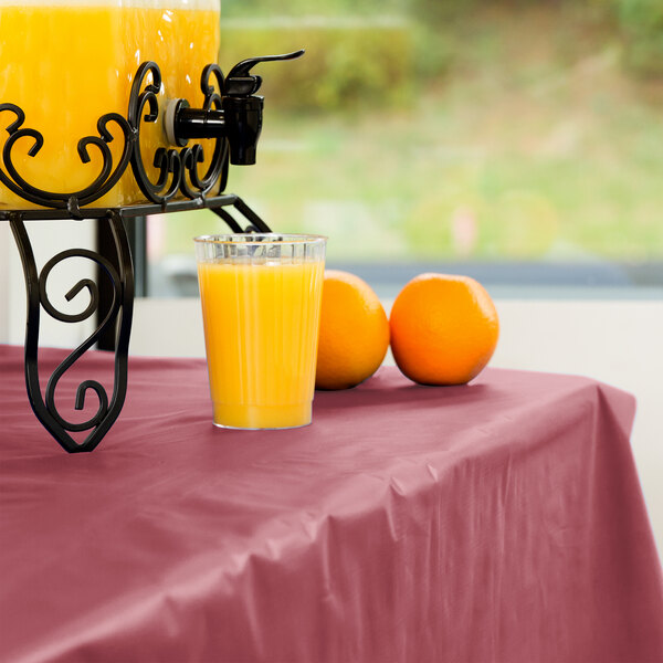 A glass of orange juice sits on a table with a Creative Converting burgundy disposable plastic tablecover.