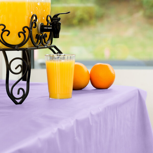 A glass of orange juice sits on a lavender plastic tablecover on a table.