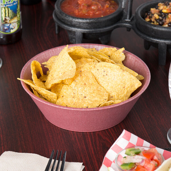 A Raspberry polyethylene round basket filled with potato chips on a table with a bowl of salsa.