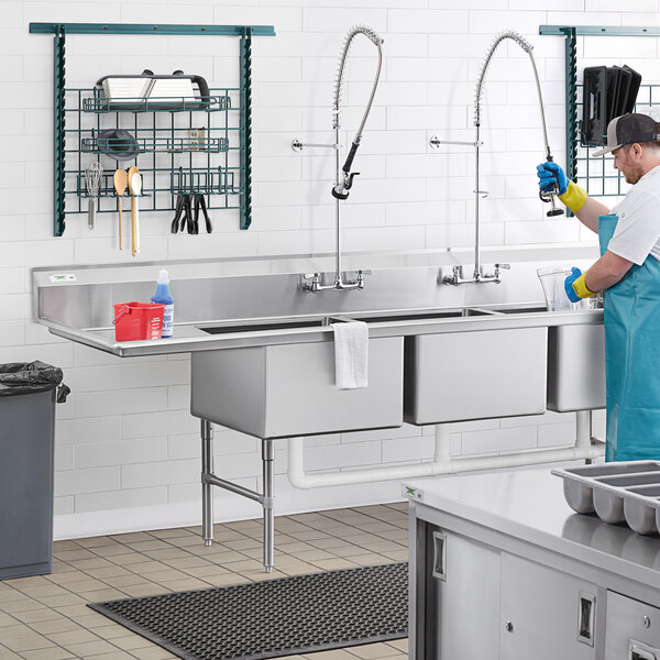 A man in a blue uniform washing dishes in a Regency stainless steel 3 compartment sink with left drainboard.