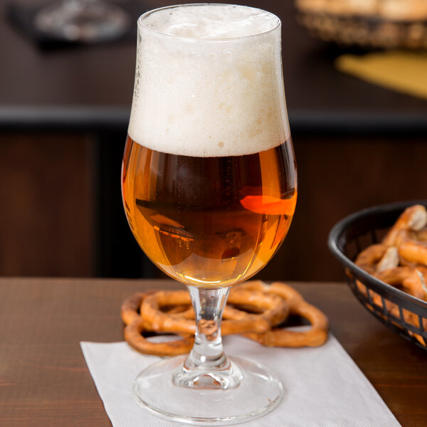A close up of a Libbey stemmed pilsner glass full of beer with foam and pretzels on the table.