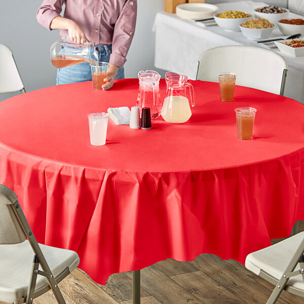 A woman sitting at an outdoor table set with a red Creative Converting plastic table cover, bowls, and glasses of liquid.