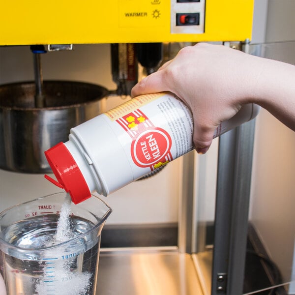 A hand pours Carnival King Kettle Kleen from a measuring cup into a container on a school kitchen counter.