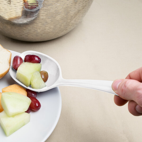 A hand holding an Elite Global Solutions white spoon filled with fruit over a bowl.