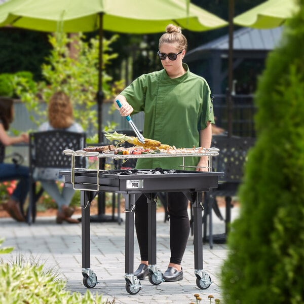 A woman in a green shirt grilling food on a Backyard Pro portable charcoal grill set up on a black table.
