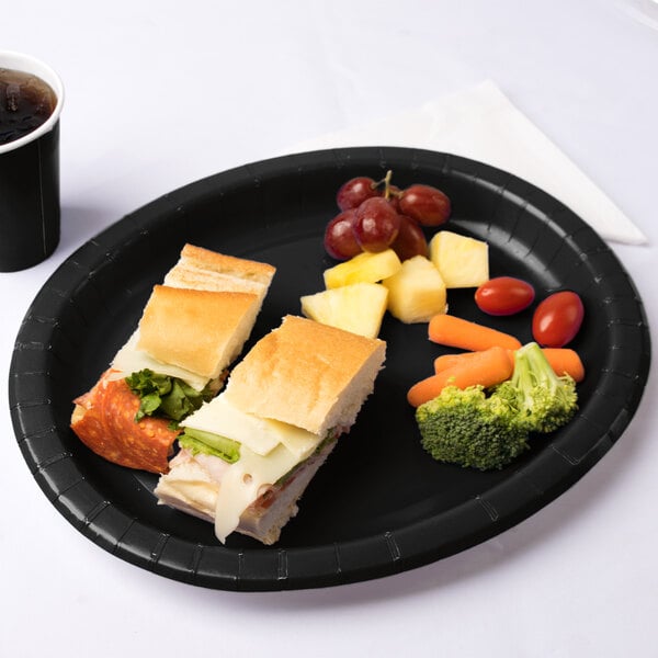A sandwich cut in half on a Creative Converting black oval paper platter with broccoli and carrots on the table.
