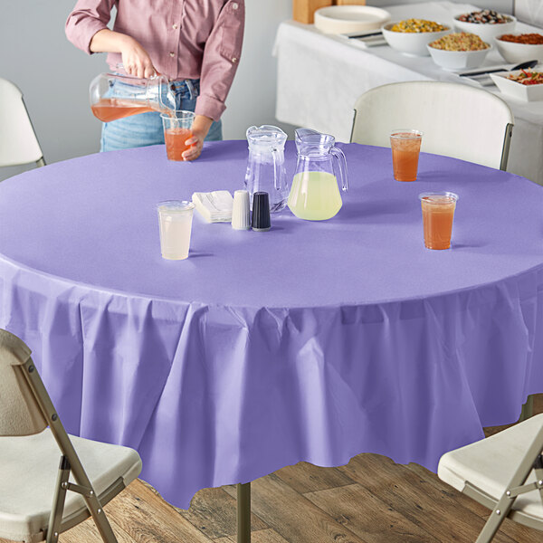 A woman pouring yellow liquid into a glass on a table with a purple Creative Converting octyround tablecloth.