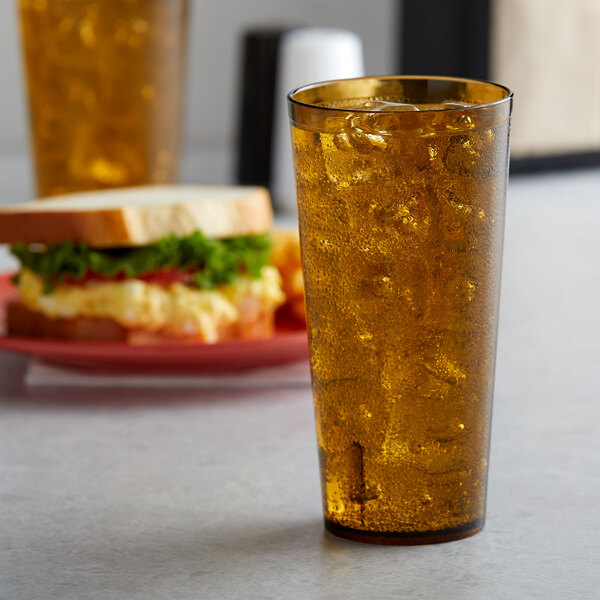 A 24 oz. amber plastic tumbler filled with a drink and ice on a table with a sandwich.