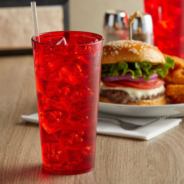 A red customizable plastic tumbler filled with red liquid and ice with a straw next to a plate of food including a burger