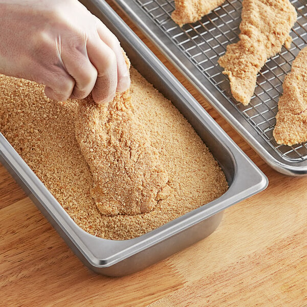A close-up of a person's hand pouring plain bread crumbs into a pan.