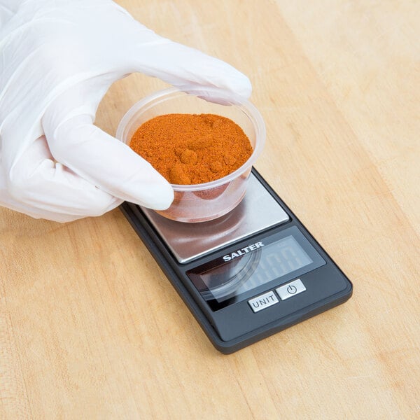 A gloved hand using a Taylor Ultra Fine Digital Scale to weigh a container of red powder.