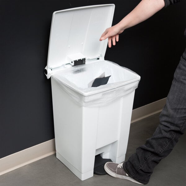 A person's hand reaching to open a white Rubbermaid step-on trash can.