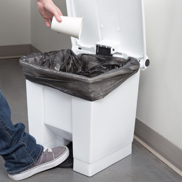 A person using a white Rubbermaid rectangular step-on trash can to put a black plastic bag into it.
