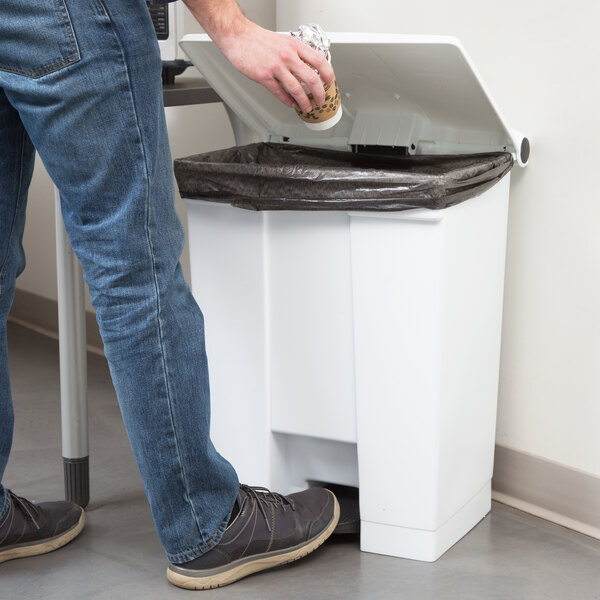 A person in jeans putting a cup into a white Rubbermaid step-on trash can.