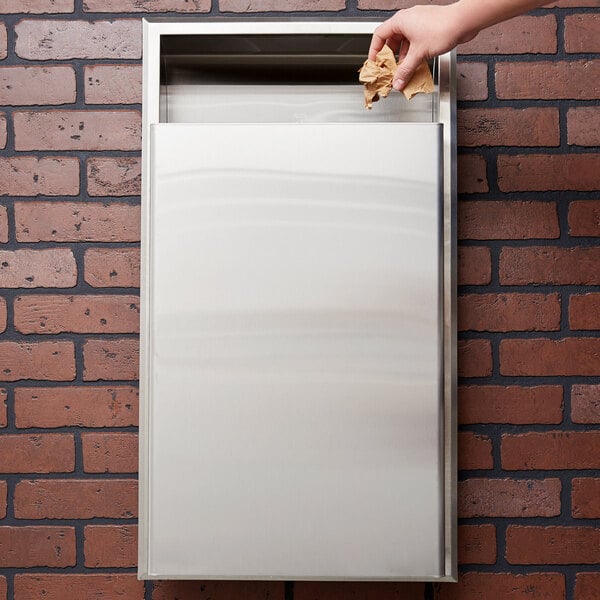 A hand throwing paper into a Bobrick recessed rectangular waste receptacle in a wall.