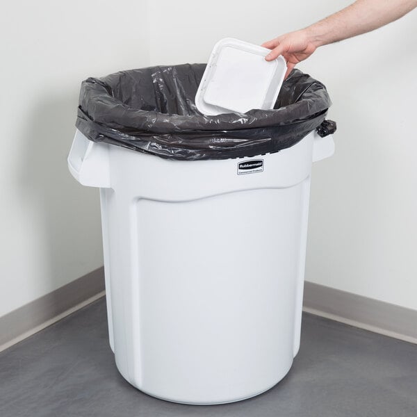 A hand placing a black plastic container in a white Rubbermaid trash can.