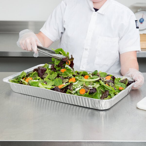 A woman in a white shirt and gloves holding a Western Plastics foil steam table pan filled with salad.