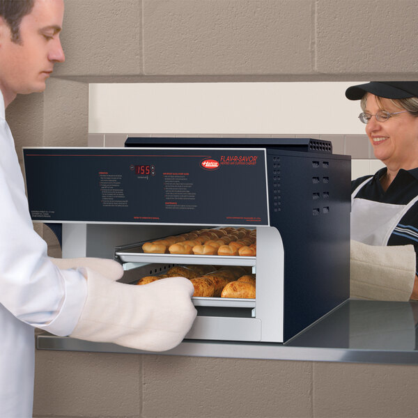 A man and woman wearing white aprons using a Hatco Flav-R-Savor pass-through heated air curtain to take bread out of an oven.