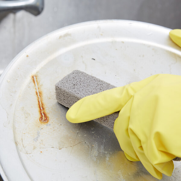 A person wearing yellow gloves uses a Pumie Heavy-Duty Scouring Stick to clean a metal pan.