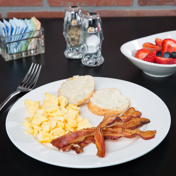 A Tuxton AlumaTux Pearl White plate with bacon, eggs, and toast with a bowl of fruit on a table.