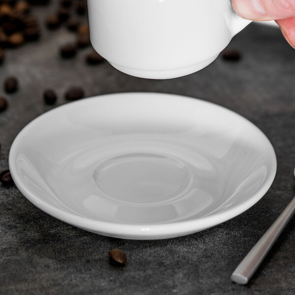A hand holding a white Tuxton saucer under a white cup.