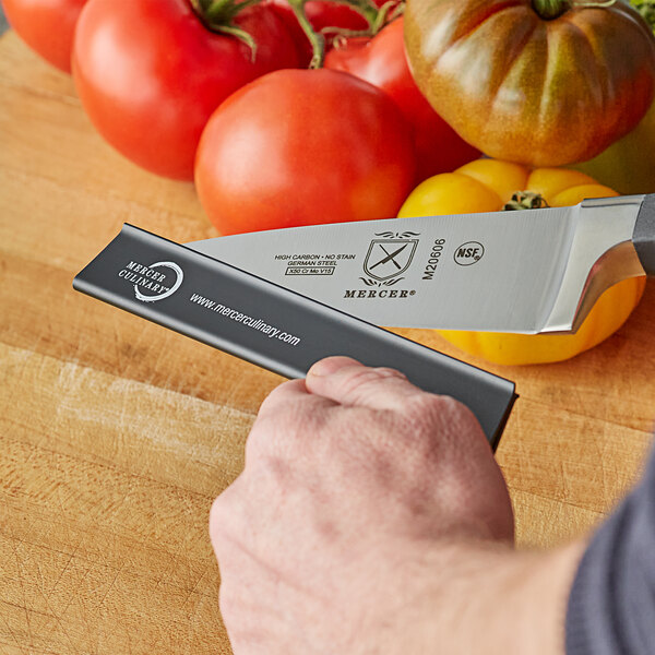 A hand holding a Mercer Culinary polypropylene knife blade guard over a knife with tomatoes on it.