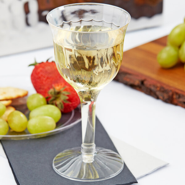 A Fineline clear plastic wine goblet filled with white wine next to a plate of grapes and a cutting board.