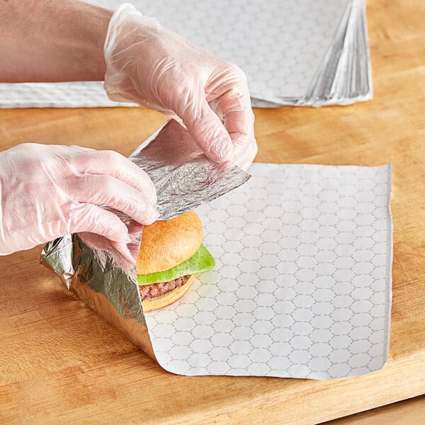 A person's hands wearing gloves wrapping a hamburger in a foil sheet.