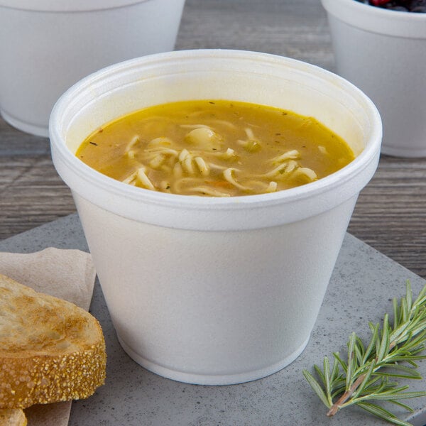 A white foam cup of soup with noodles and a piece of bread on a table.