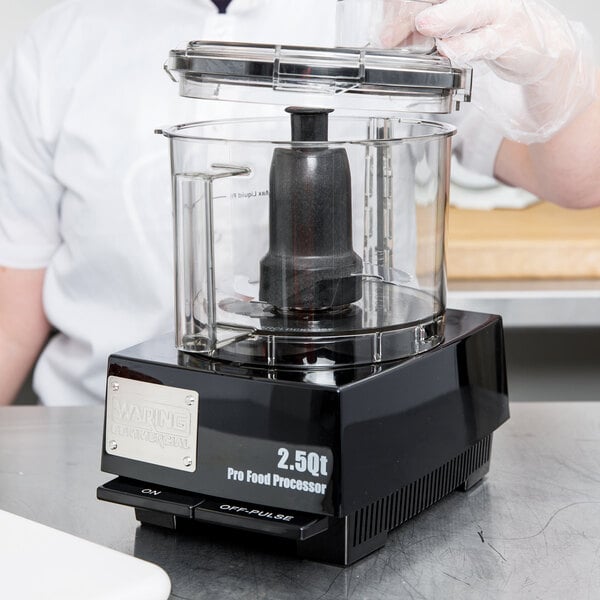 A person in a white coat and gloves using a Waring commercial food processor on a counter.