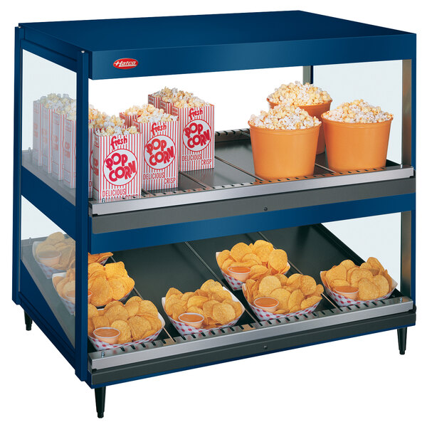 A blue Hatco countertop display case with shelves of popcorn and potato chips.