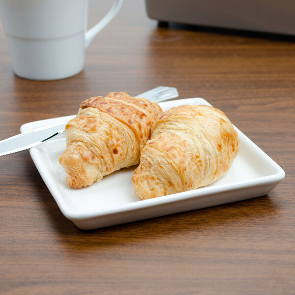 A Tuxton AlumaTux Pearl White square china plate with two croissants and a knife on it.