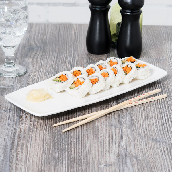 A rectangular white china plate with sushi and chopsticks on a wood table.