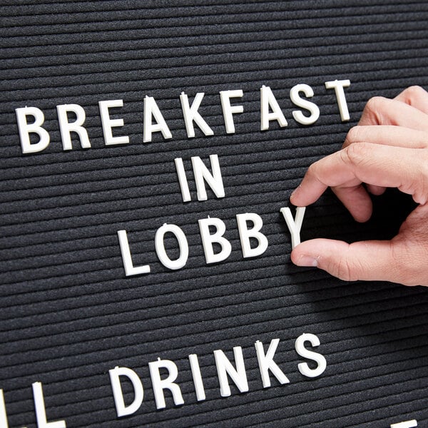 A hand touches a black sign with white Aarco Helvetica lettering that spells "breakfast in lobby".